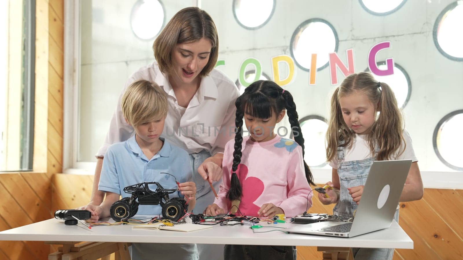 Teacher teach and explain students about digital electrical tool or car model. Diverse kid learn electronic equipment and use to fix motherboard or robotic model at table with wires placed. Erudition.