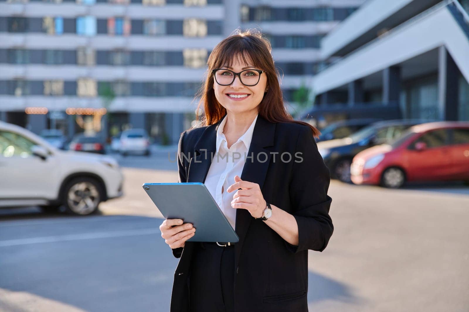 Mature confident successful business woman with digital tablet in black suit looking at camera outdoors, backdrop of modern city. Business, entrepreneurship mentoring insurance sales advertising work