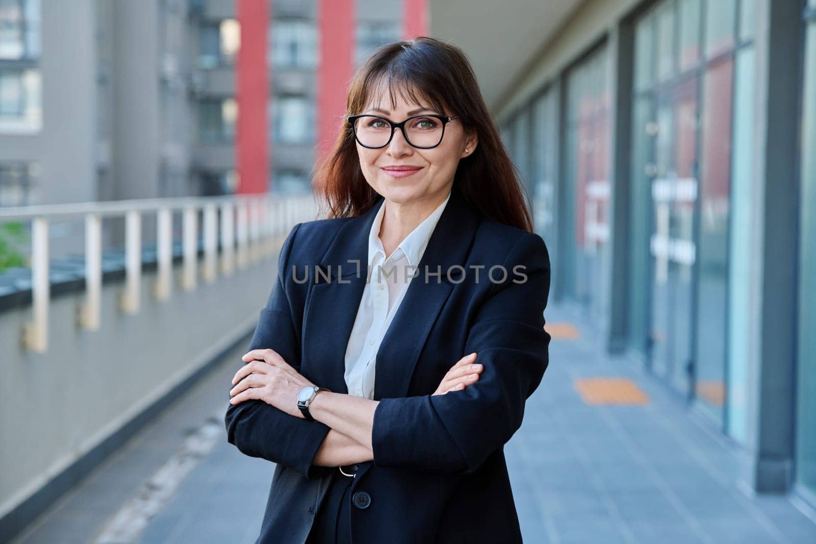 Mature confident successful business woman with crossed arms in black suit looking at camera outdoors, backdrop of modern city. Business, entrepreneurship mentoring insurance sales advertising work