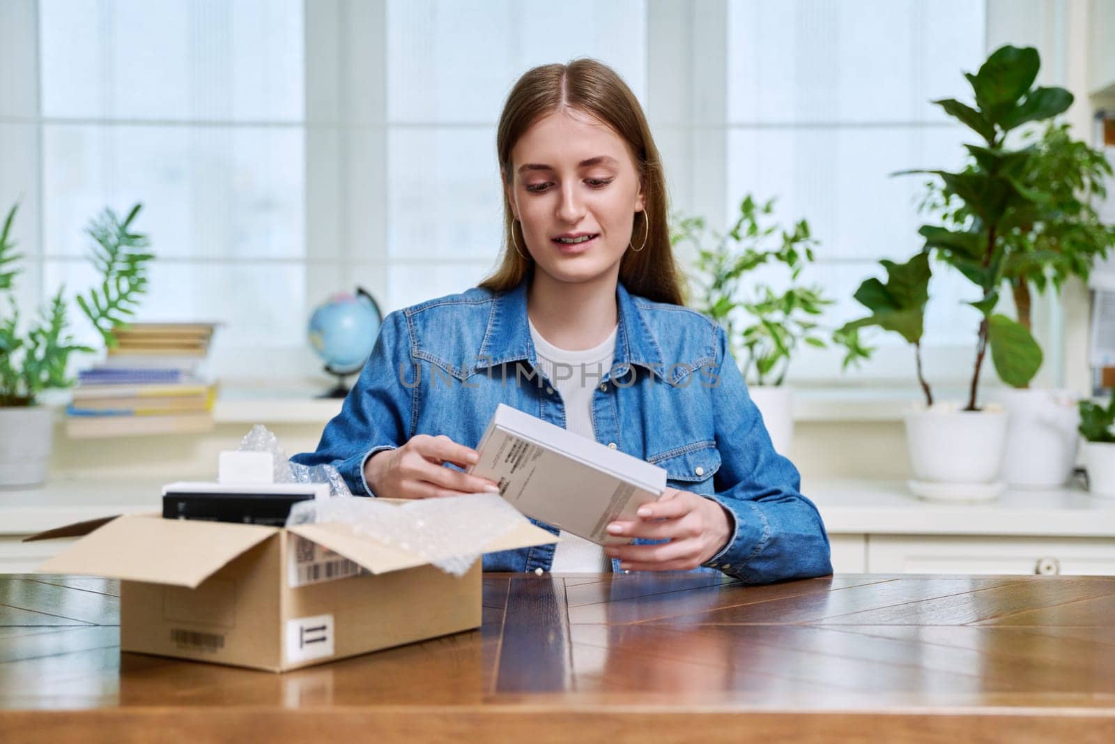 Satisfied young teen female shopper consumer sits at home and unpacks cardboard box with online purchases. Teenager girl unpacking box with new smartphone, headphones. Delivery by mail, online store