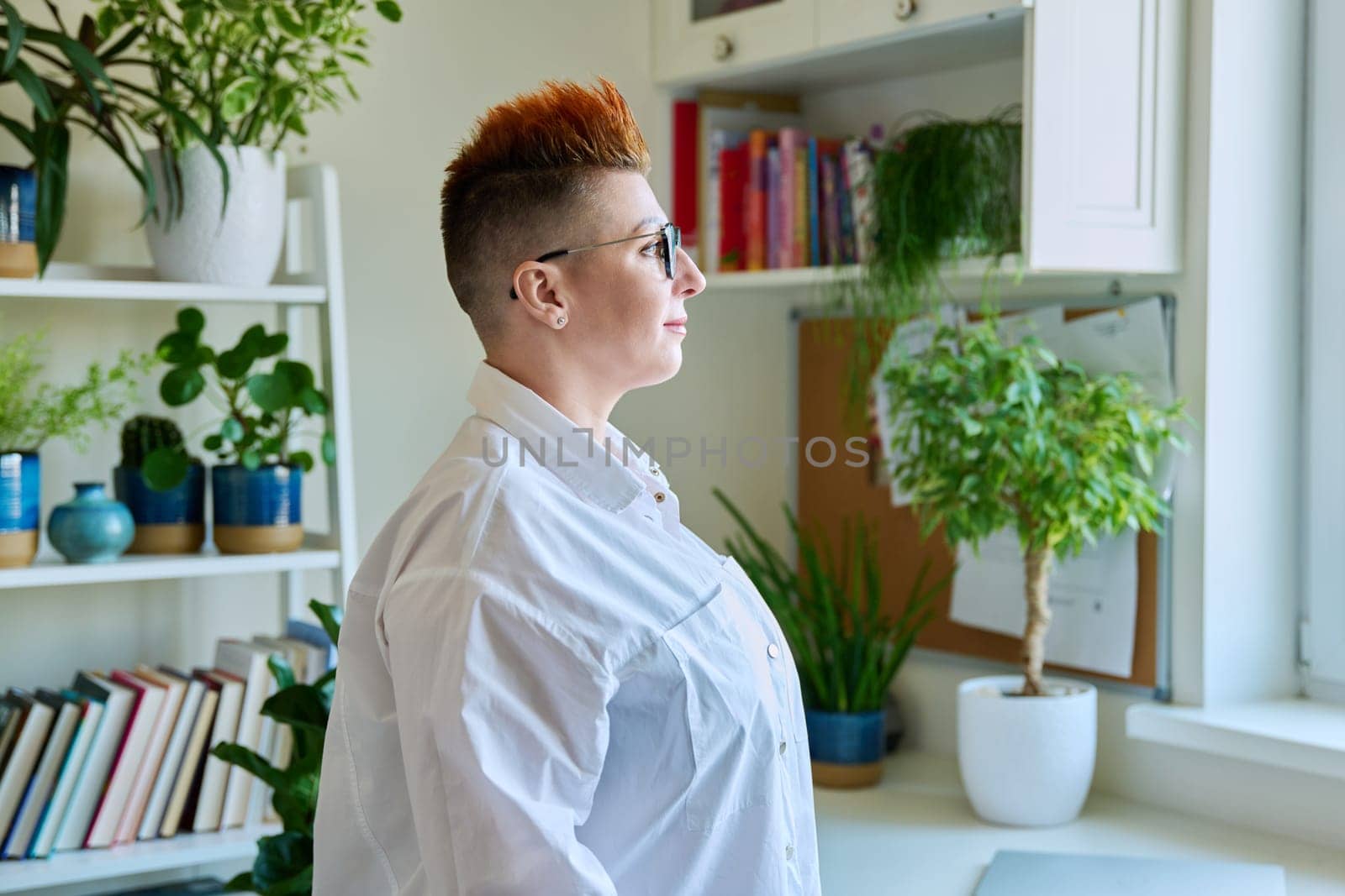 Profile portrait of serious middle-aged woman in glasses with red haircut looking at window in home interior, copy space. Mature people, lifestyle, health, life concept