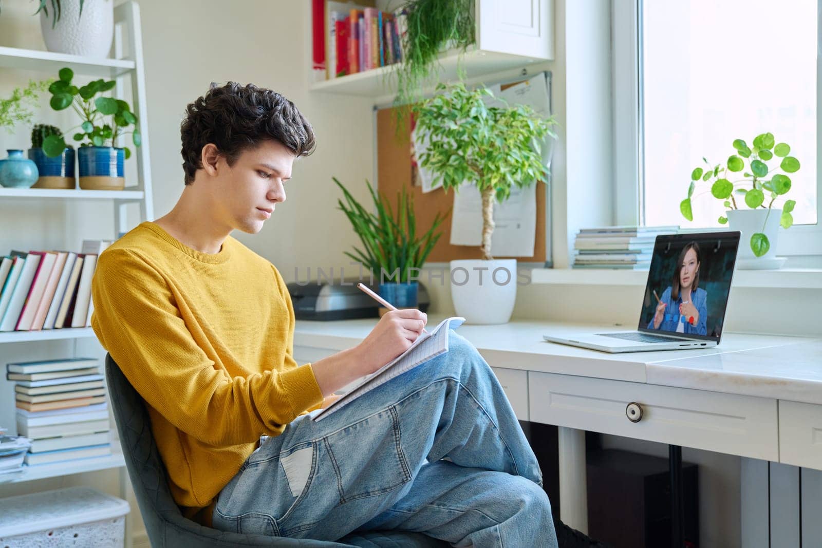 Guy college student sitting at home at desk looking at laptop screen, making notes in notebook. Young male studying online, watching webinar, browsing Internet. E-learning, education, technology youth