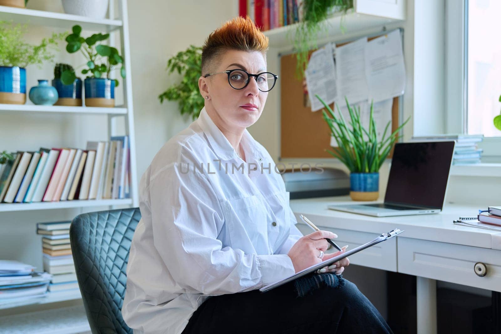 Middle aged confident successful business woman with clipboard sitting at workplace. Remote business, technologies, internet, teaching, blog, work concept