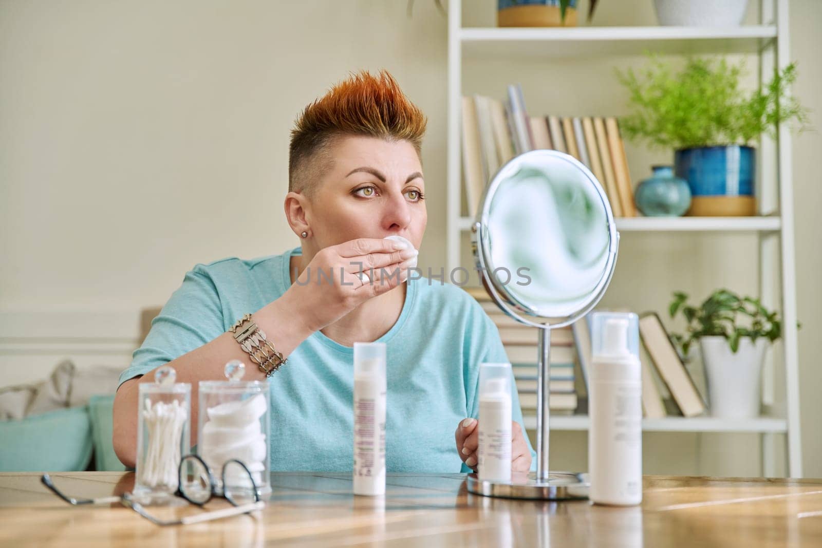 Middle-aged woman with a mirror taking care of her facial skin by VH-studio