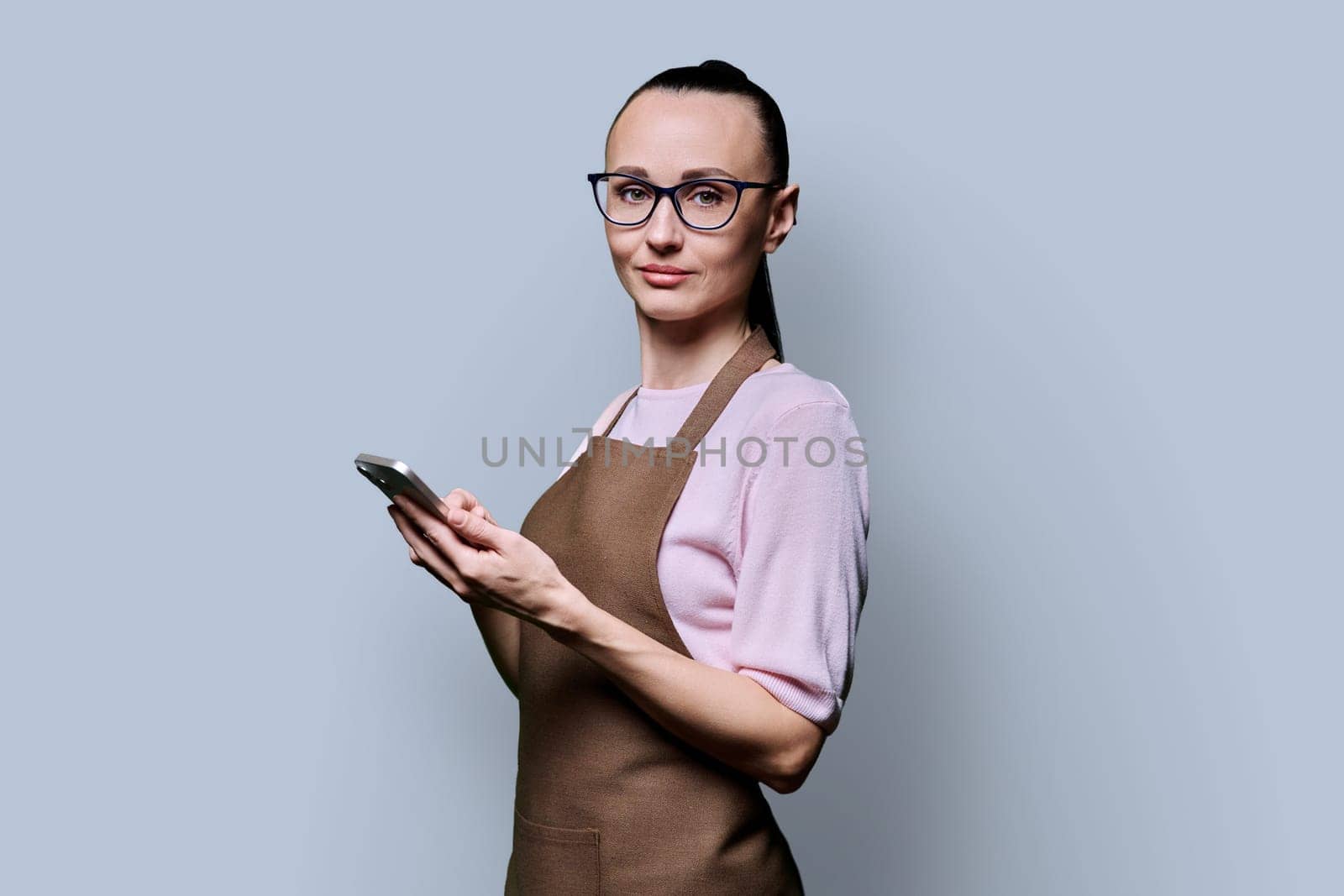 Portrait of 30s woman in apron with smartphone looking at camera on grey background. Serious female using mobile phone texting receiving sending order. Technologies applications service small business