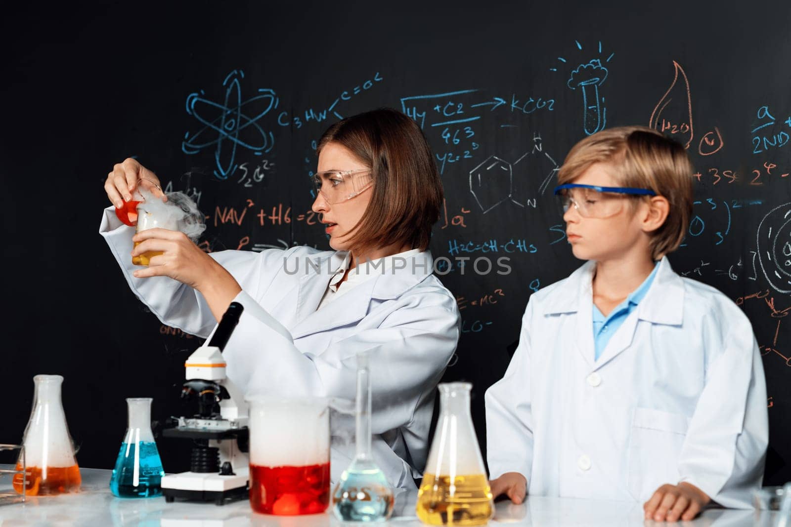 Skilled teacher and student in laboratory wear lab coat stand and experiment about science of chemistry in STEM class. Teacher pour red liquid down to yellow beaker. Table put many flask. Erudition.