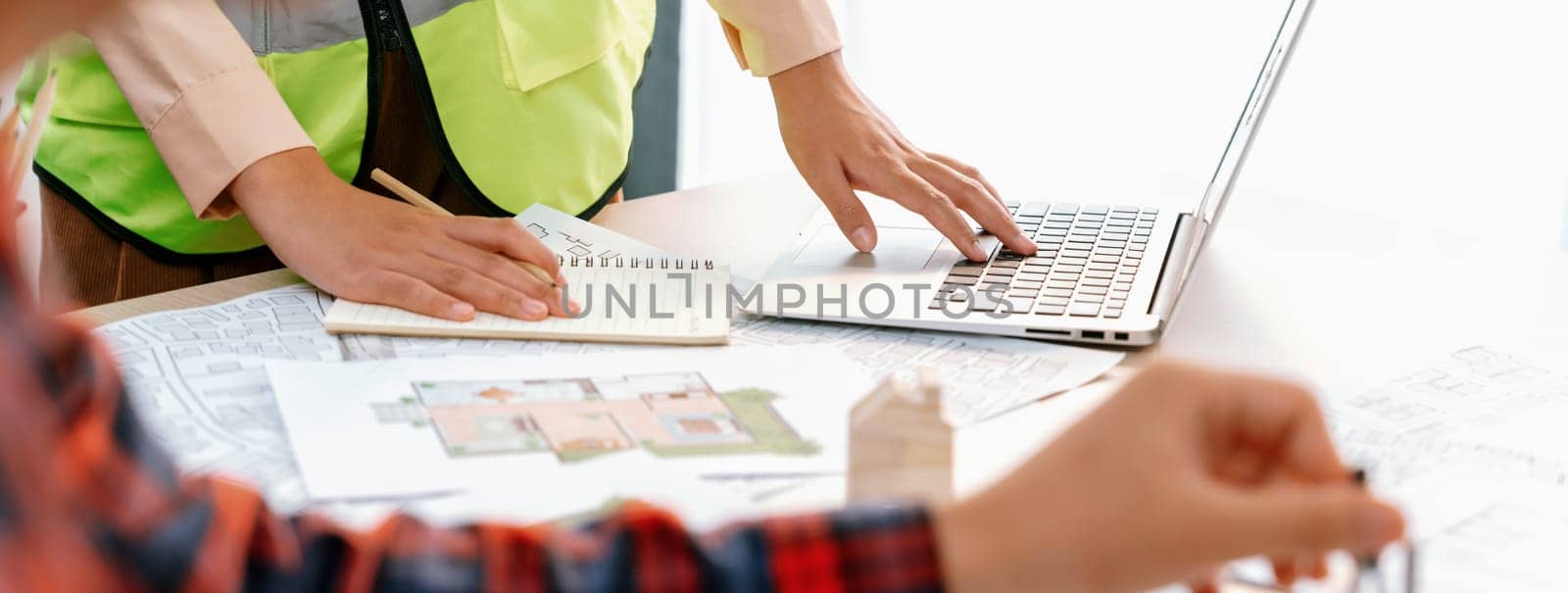 Cropped image of professional engineer team working on blueprint while his coworker working on laptop at meeting table with blueprint and wooden block scattered around. Closeup. Delineation.