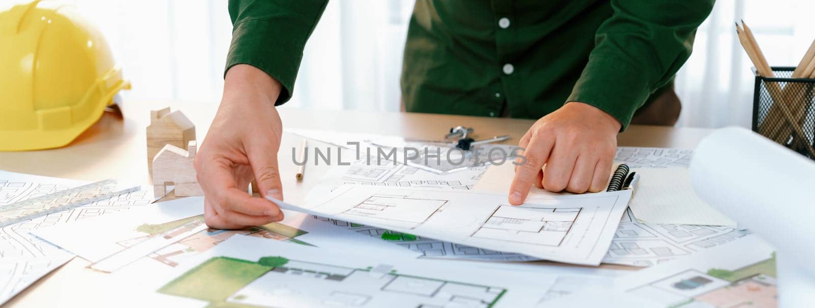 A portrait of professional male engineer shows mistake point in blueprint on messy table with architectural plan and yellow helmet placed at modern office. Closeup. Focus on hand. Delineation.
