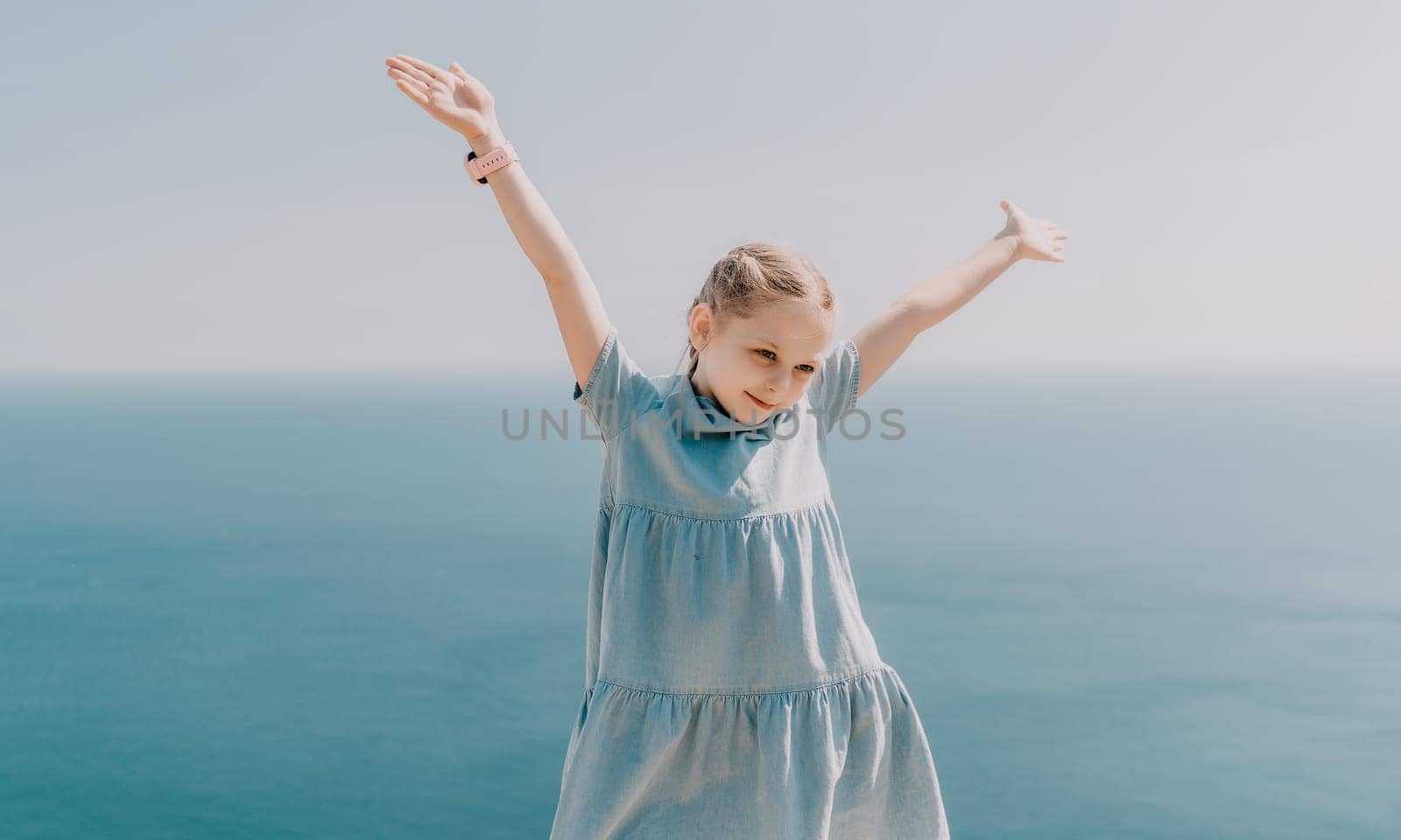 A young girl is standing on a beach, wearing a blue dress and holding her arms up in the air. Concept of joy and freedom, as the girl appears to be enjoying her time by the ocean