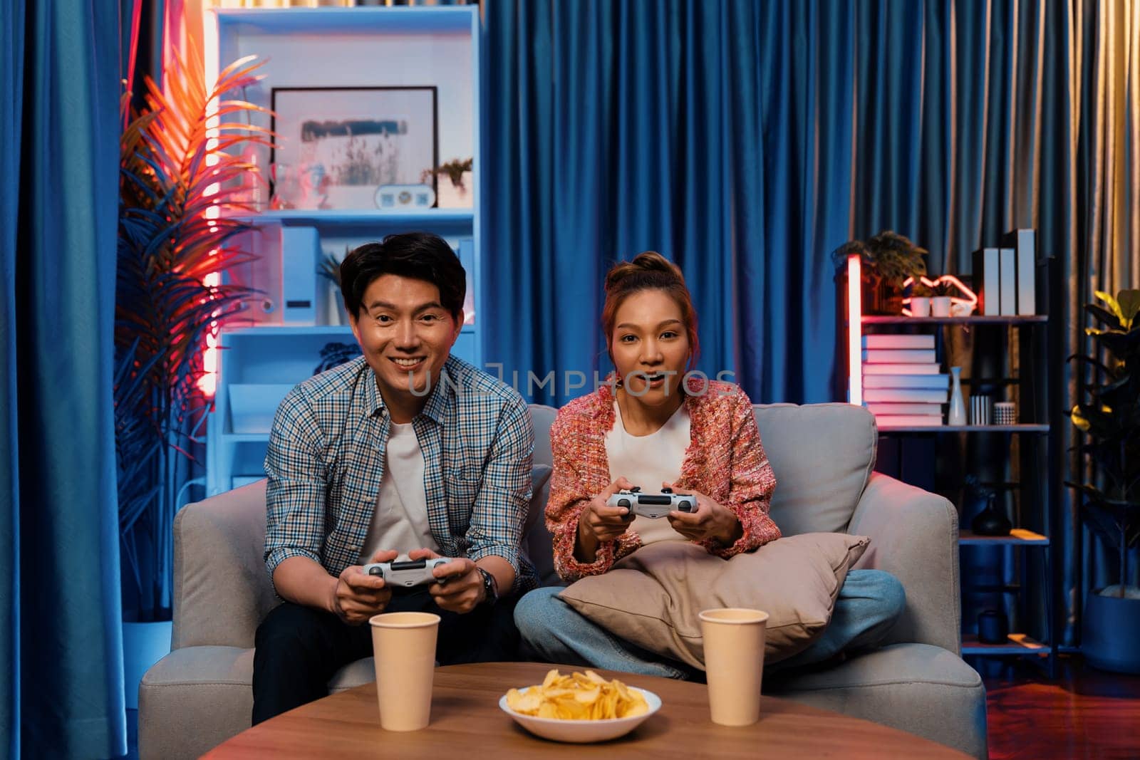 Sitting sofa of happy smiling couple playing video game control by joystick fronted snack and drinks competing challenge level with funny at modern neon blub light comfy living room at home. Infobahn.