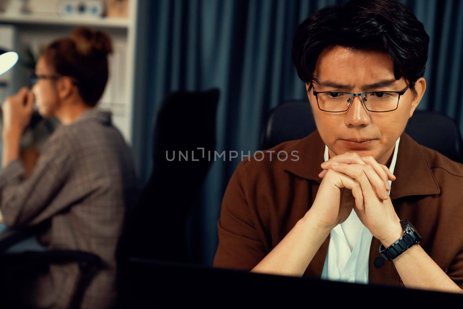Businessman with stressful face working on laptop waiting email while coworker trading stock exchange investment at late night time at modern office, working on startup company project job. Infobahn.