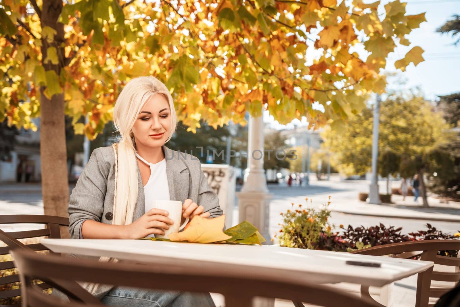 A blonde woman sits at a table with a cup of coffee and a leaf on it. The scene is set in a city with a tree in the background. by Matiunina