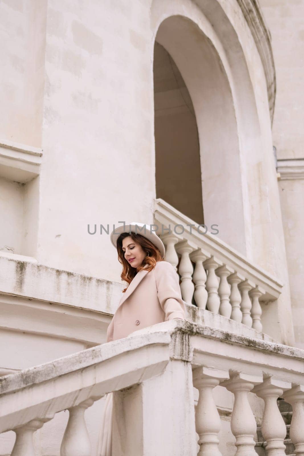 woman in elegant coat and hat against an intricate architectural backdrop, harmoniously blending modern fashion with historical allure. The soft daylight adds to its timeless appeal. by Matiunina