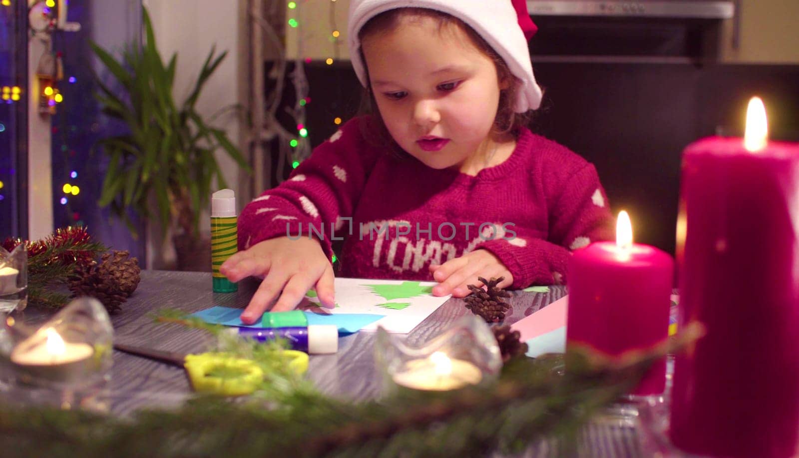 Children handicraft. Little girl glues New Year card. Table with Christmas decorations. French text on a sweater - falls