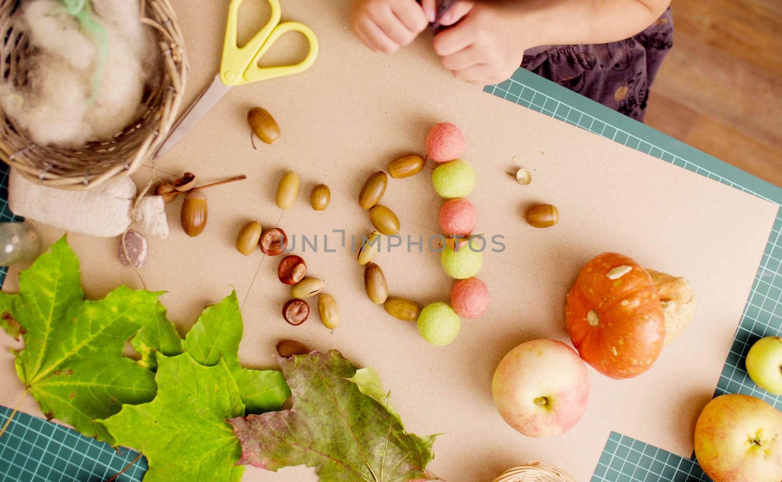 Top view. Autumn. Little girl sitting at the desk makes necklace of acorns and colored balls