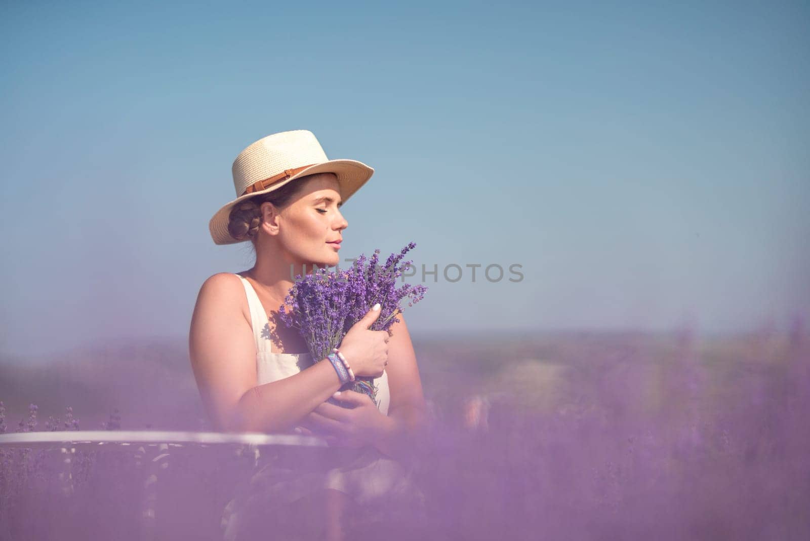 A woman is sitting in a field of lavender flowers and wearing a straw hat. She is smiling and holding a bouquet of flowers. Scene is peaceful and serene, as the woman is surrounded by the beauty of nature.