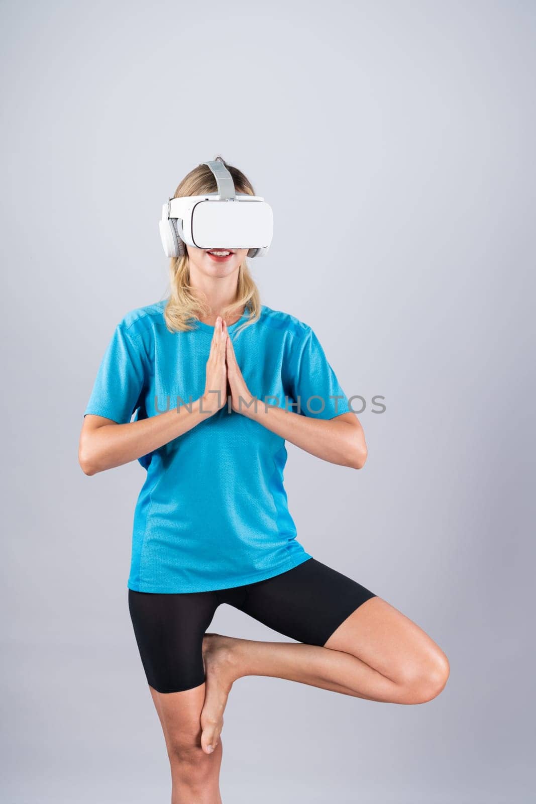 Caucasian girl practicing yoga while wearing shirt with pink background. Happy smart woman making yoga posing while standing at pink studio and using VR goggles or visual reality headset. Contraption.