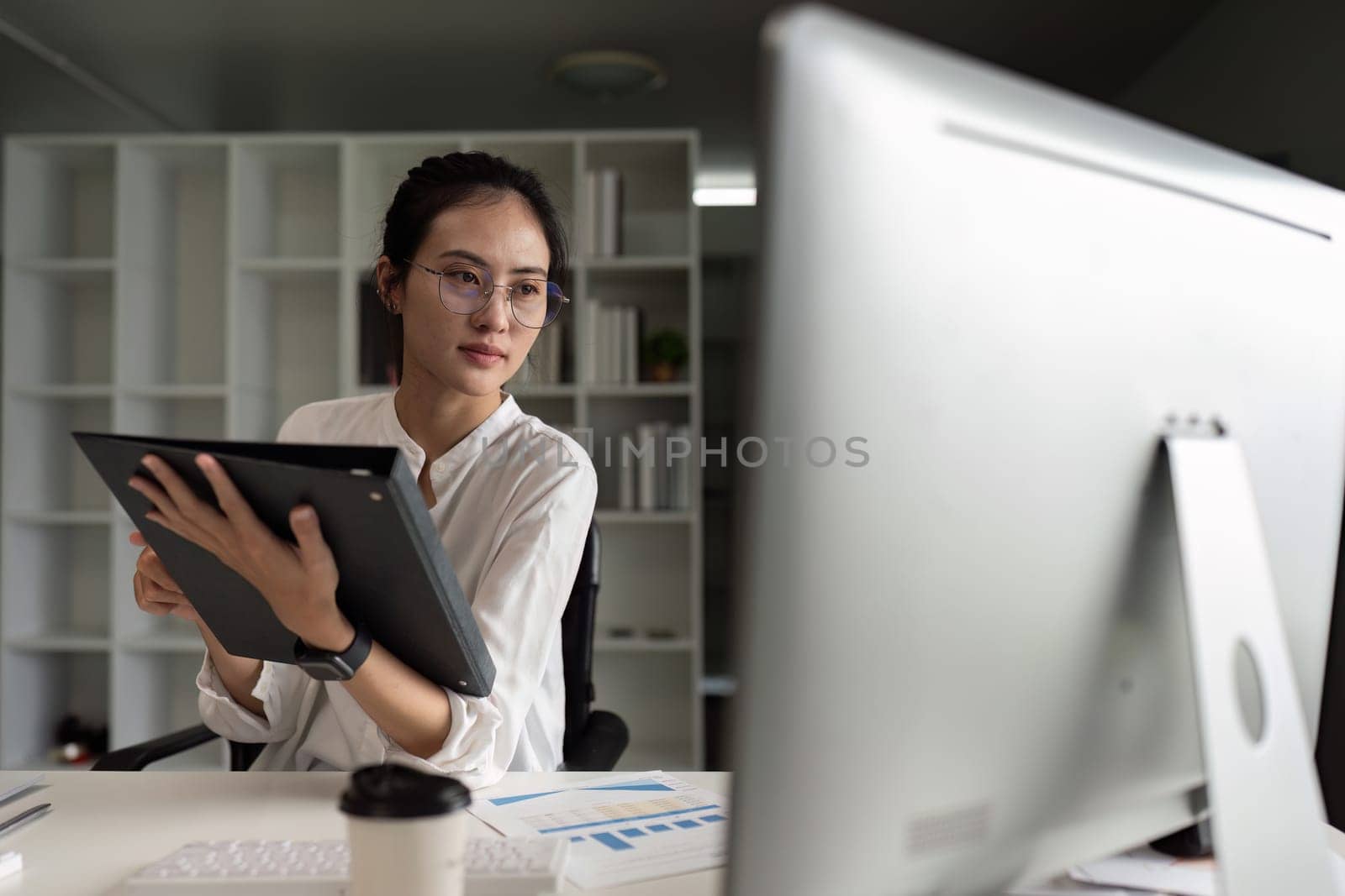 A woman is sitting at a desk with a computer monitor in front of her. She is holding a folder in her hand and she is focused on the task at hand. Concept of productivity and concentration