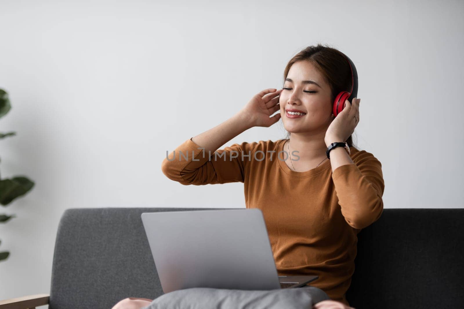Young Asian woman spends her free time at home sitting on the sofa wearing headphones listening to music to relax and have fun. by wichayada