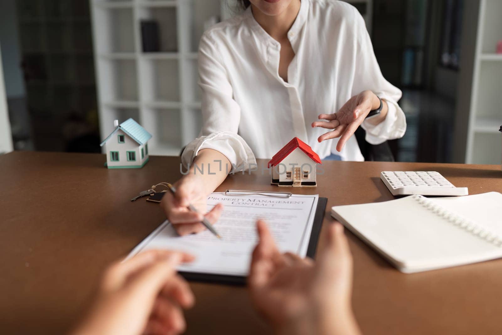 A woman is signing a document in front of a house model. The woman is wearing a white shirt and the house model is red