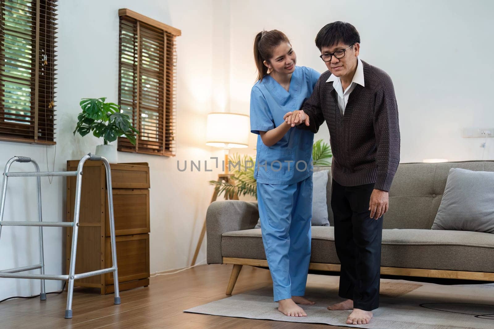 A woman in a blue uniform helps an older man with a walker. The scene is set in a living room with a couch and a potted plant. Scene is caring and supportive