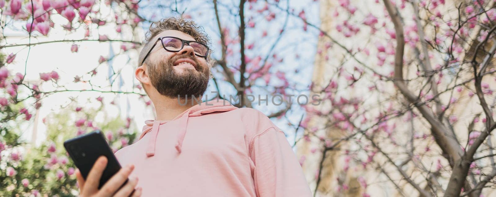 Banner Spring day. Bearded man in pink shirt talking by phone. Spring pink sakura blossom. Handsome young man with smartphone. Fashionable man in trendy glasses. Bearded stylish man. Copy space by Satura86