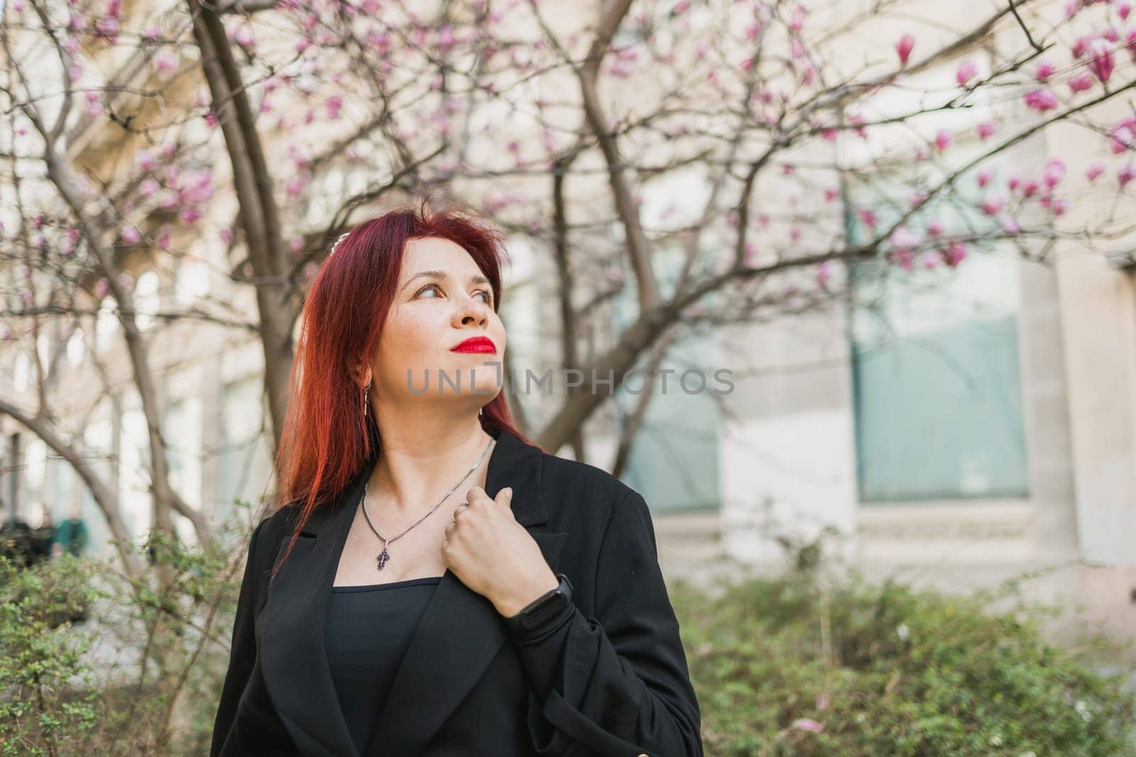 Fashion outdoor photo of beautiful woman with red curly hair in elegant suit posing in spring flowering park with magnolias tree. Copy space and empty place for advertising text.