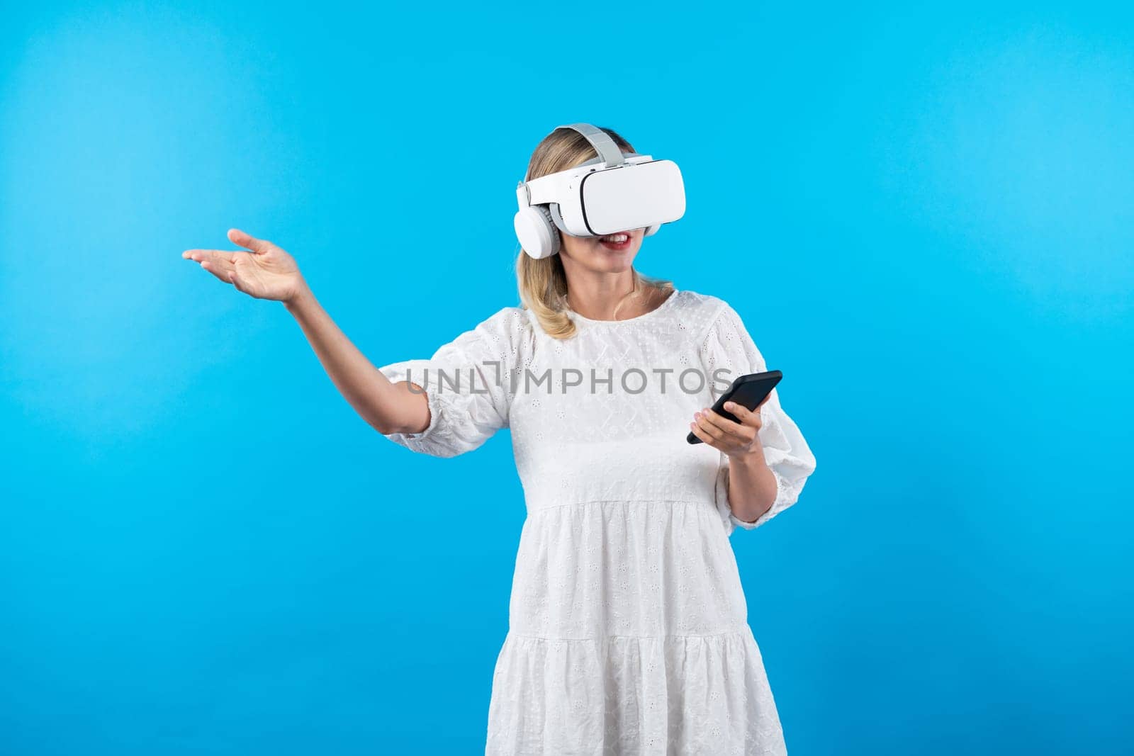 Caucasian girl wearing VR glasses while holding phone and pointing at view. Happy woman surprised while looking around to explore visual reality world or metaverse. Technology innovation. Contraption.