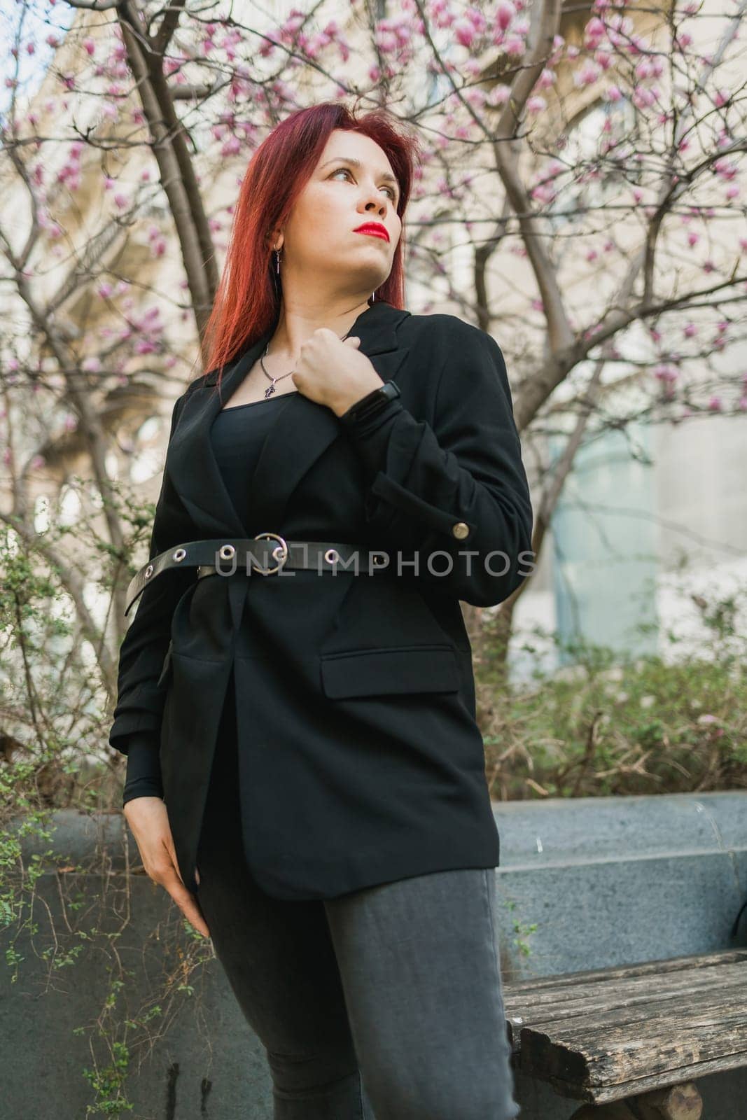 Fashion outdoor photo of beautiful woman with red curly hair in elegant suit posing in spring flowering park with magnolias tree. Copy space and empty place for advertising text by Satura86