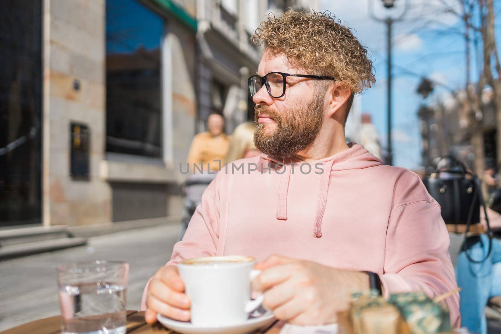 Young bearded man having breakfast at table of street cafe on spring day, drinking warm cappuccino and enjoy morning. by Satura86