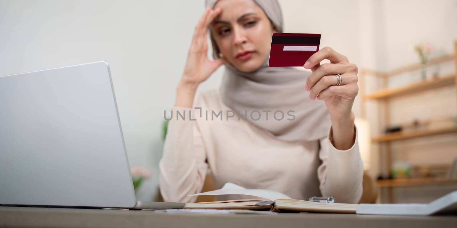 Credit card problem. Worried and stress young muslim woman in hijab sitting in living room working and make purchase in online store on laptop.