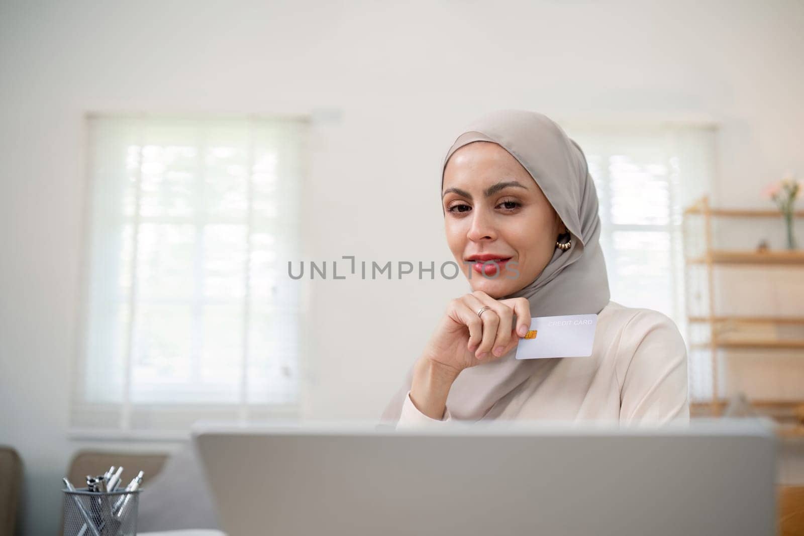 Young happy muslim woman in hijab at home using laptop shopping online with credit card while sitting on desk by nateemee