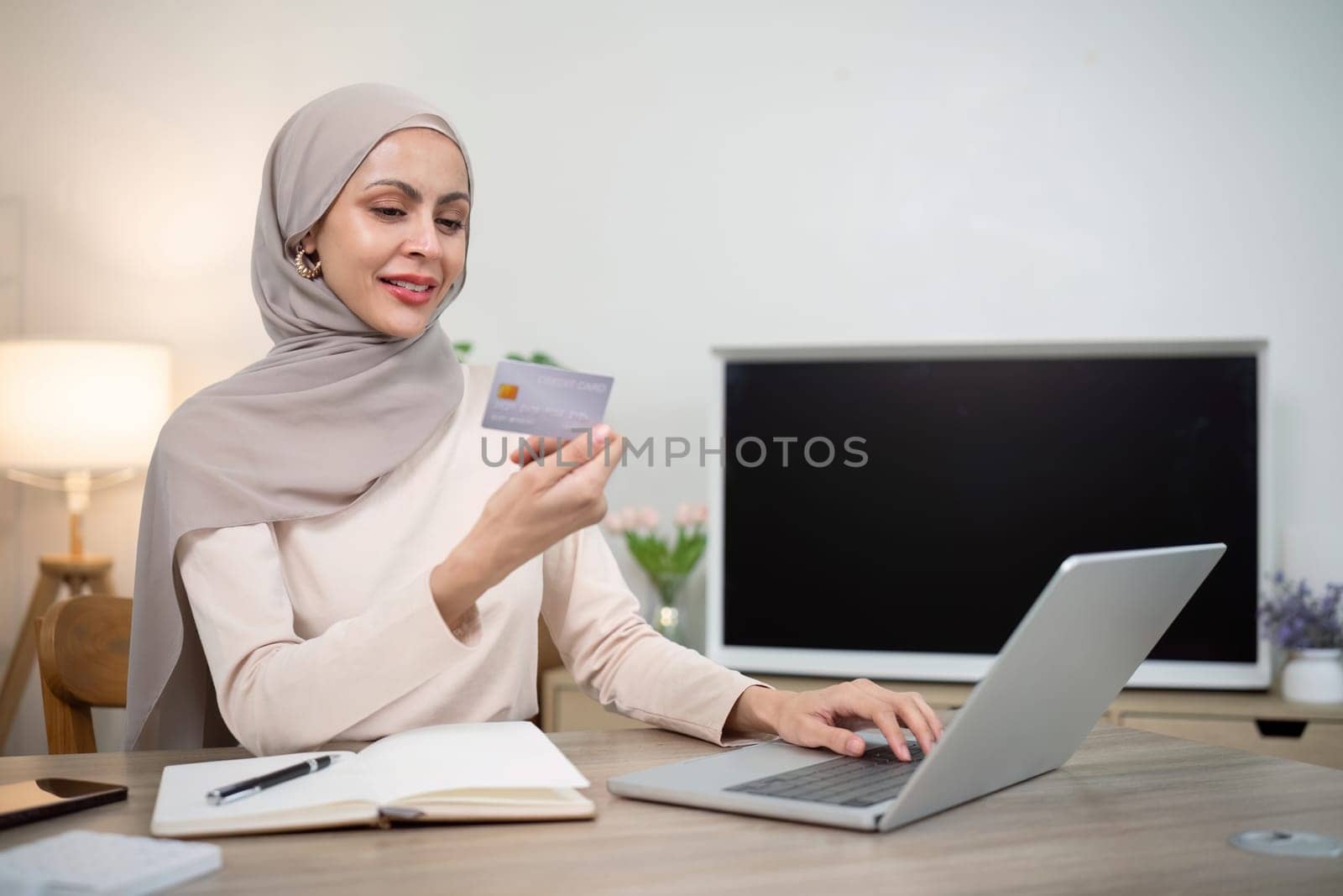 Young happy muslim woman in hijab at home using laptop shopping online with credit card while sitting on desk by nateemee