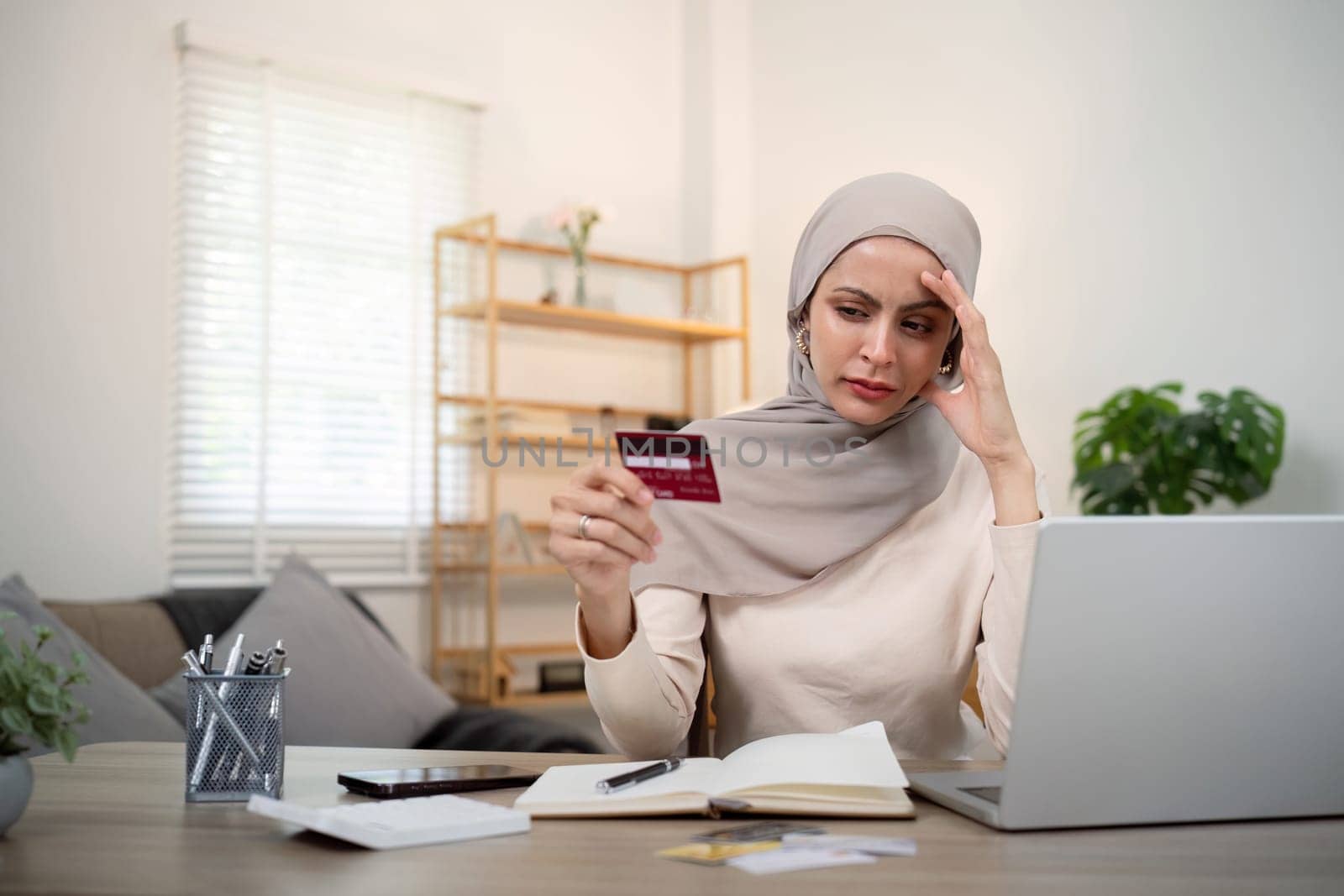 Credit card problem. Worried and stress young muslim woman in hijab sitting in living room working and make purchase in online store on laptop by nateemee