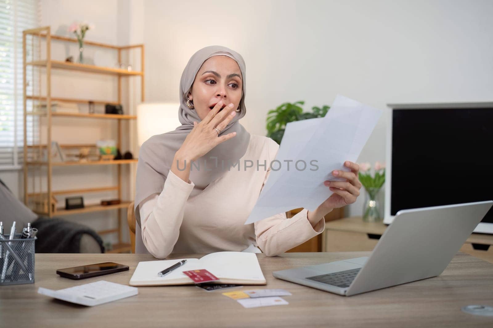 Young worried and shocked Muslim woman in hijab holding documents and calculator. Financial problems with accounts, loans, family budget at the home by nateemee