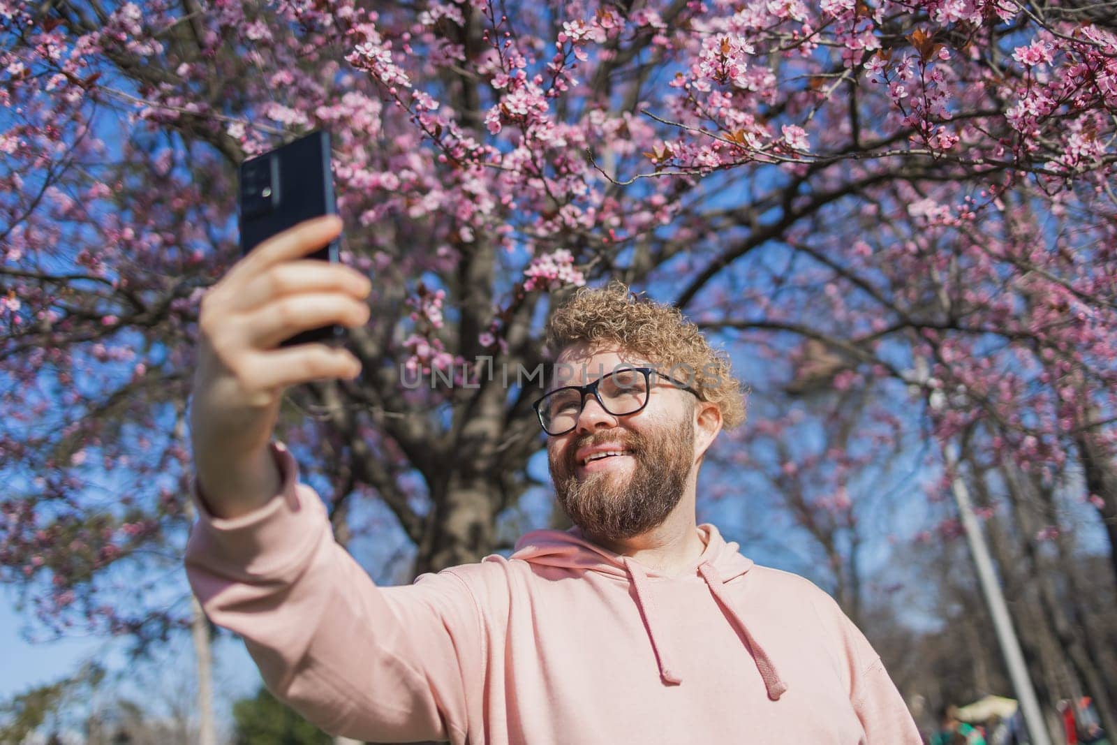 Happy curly man takes selfie against backdrop of flowering tree in spring for his internet communications. Weekend and social networks