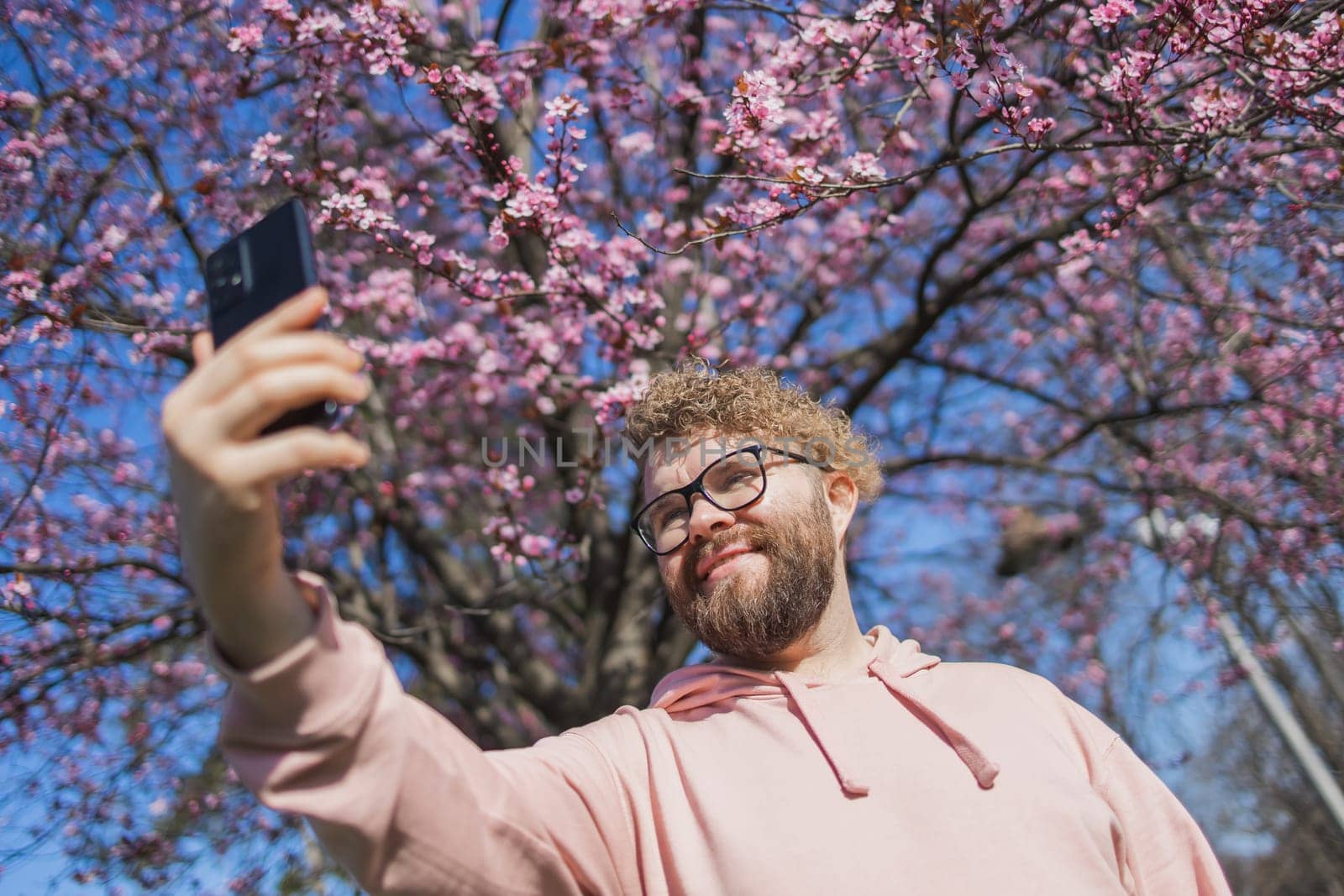 Spring day. Bearded man in pink shirt talking by phone. Spring pink sakura blossom. Handsome young man with smartphone. Fashionable man in trendy glasses. Bearded stylish man. Copy space by Satura86