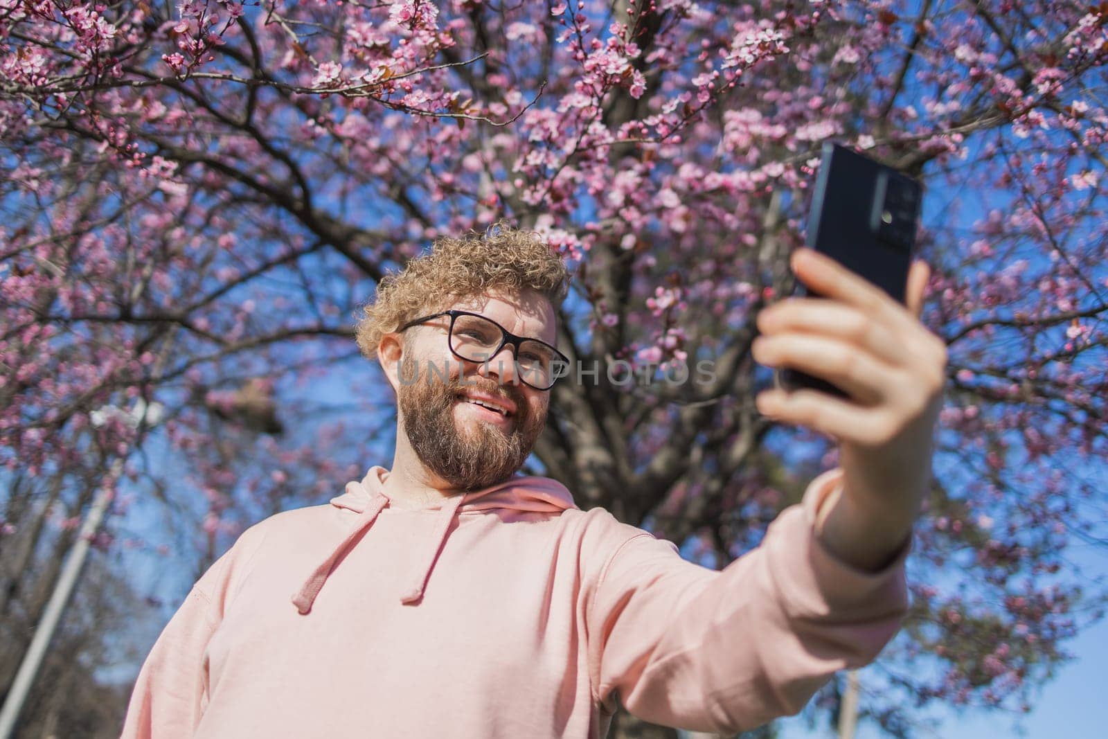 Happy curly man takes selfie against backdrop of flowering tree in spring for his internet communications. Weekend and social networks concept by Satura86