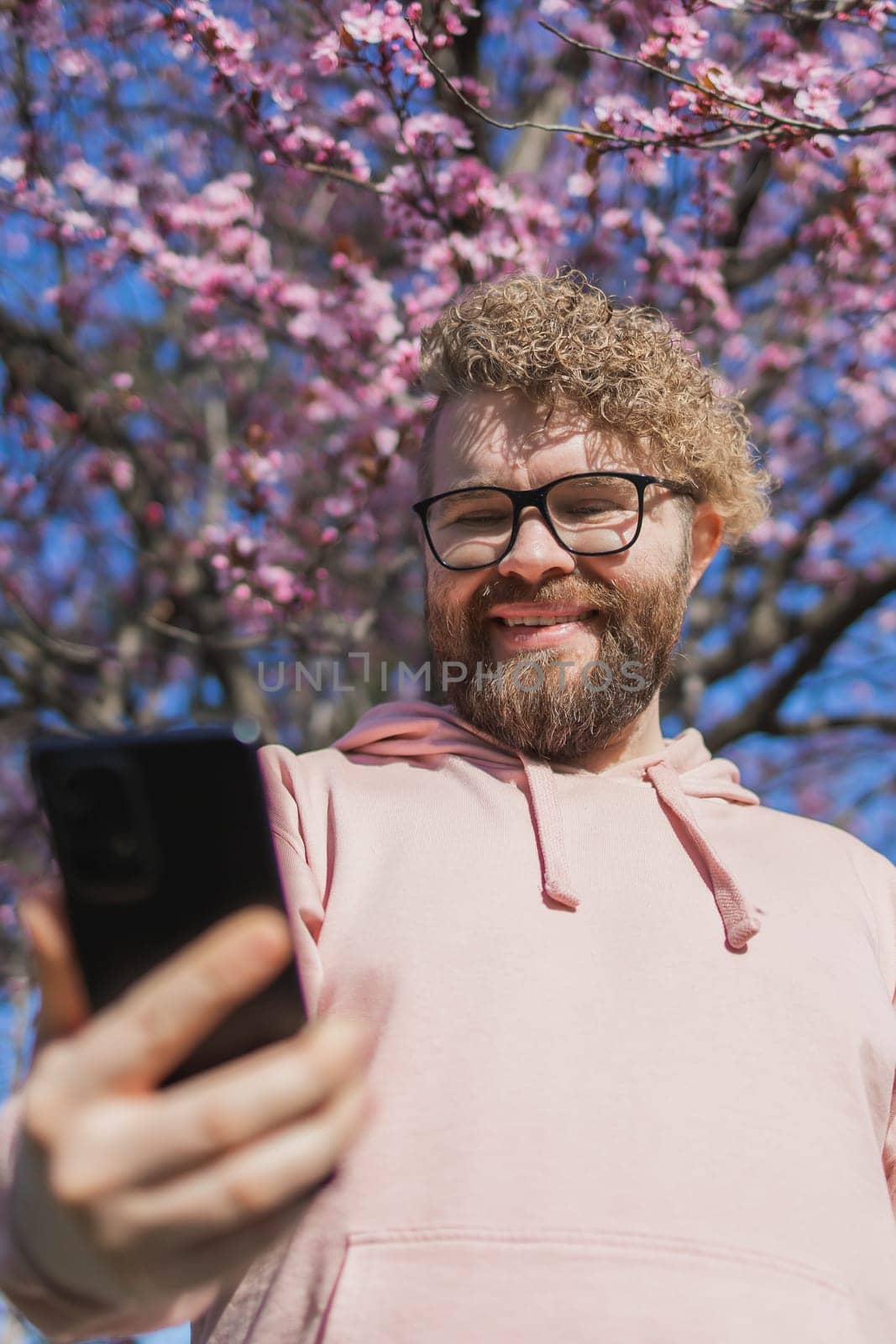 Spring day. Bearded man in pink shirt talking by phone. Spring pink sakura blossom. Handsome young man with smartphone. Fashionable man in trendy glasses. Bearded stylish man. Copy space by Satura86