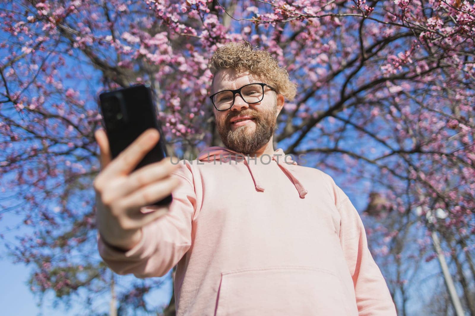 Spring day. Bearded man in pink shirt talking by phone. Spring pink sakura blossom. Handsome young man with smartphone. Fashionable man in trendy glasses. Bearded stylish man. Copy space by Satura86