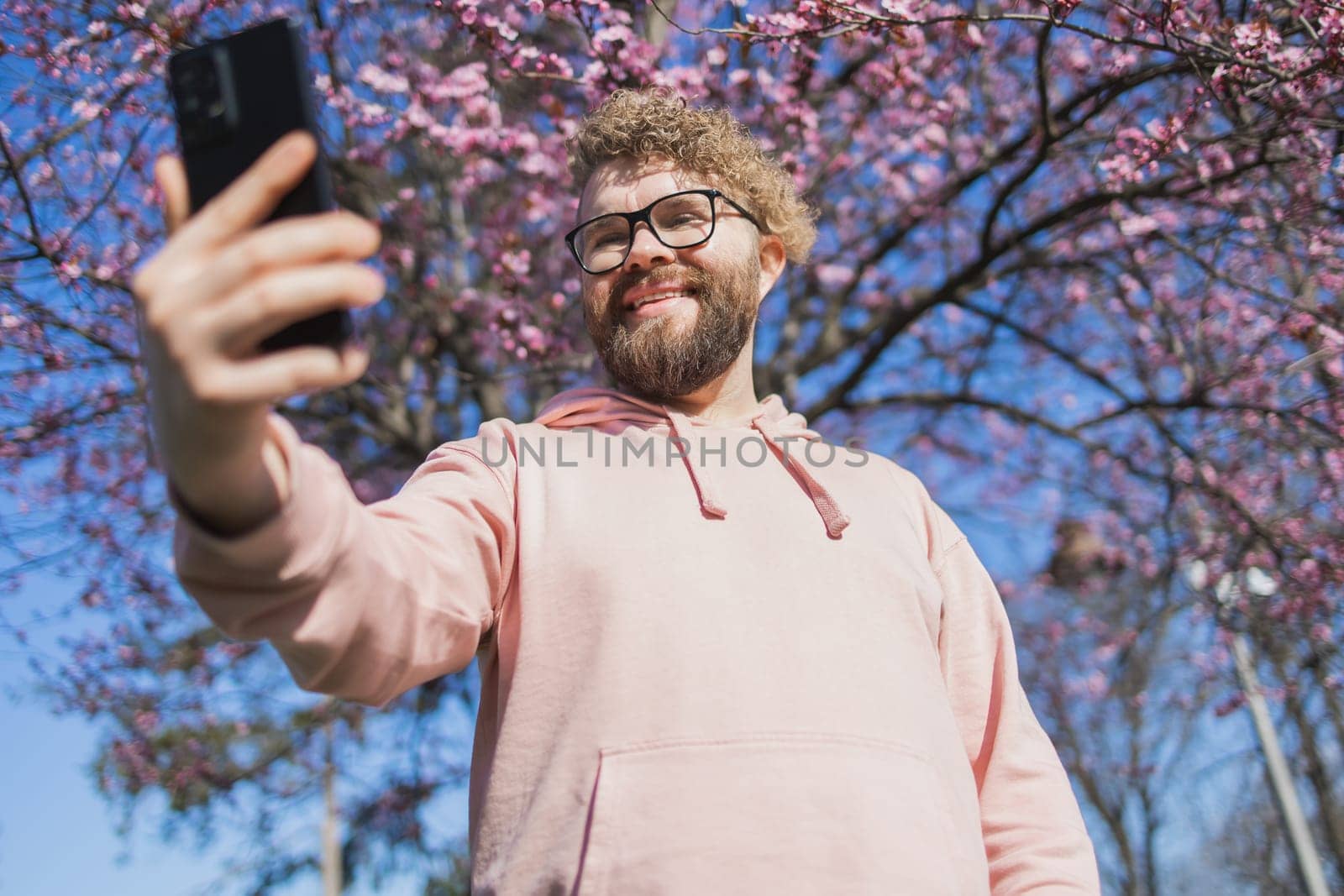 Happy curly man takes selfie against backdrop of flowering tree in spring for his internet communications. Weekend and social networks