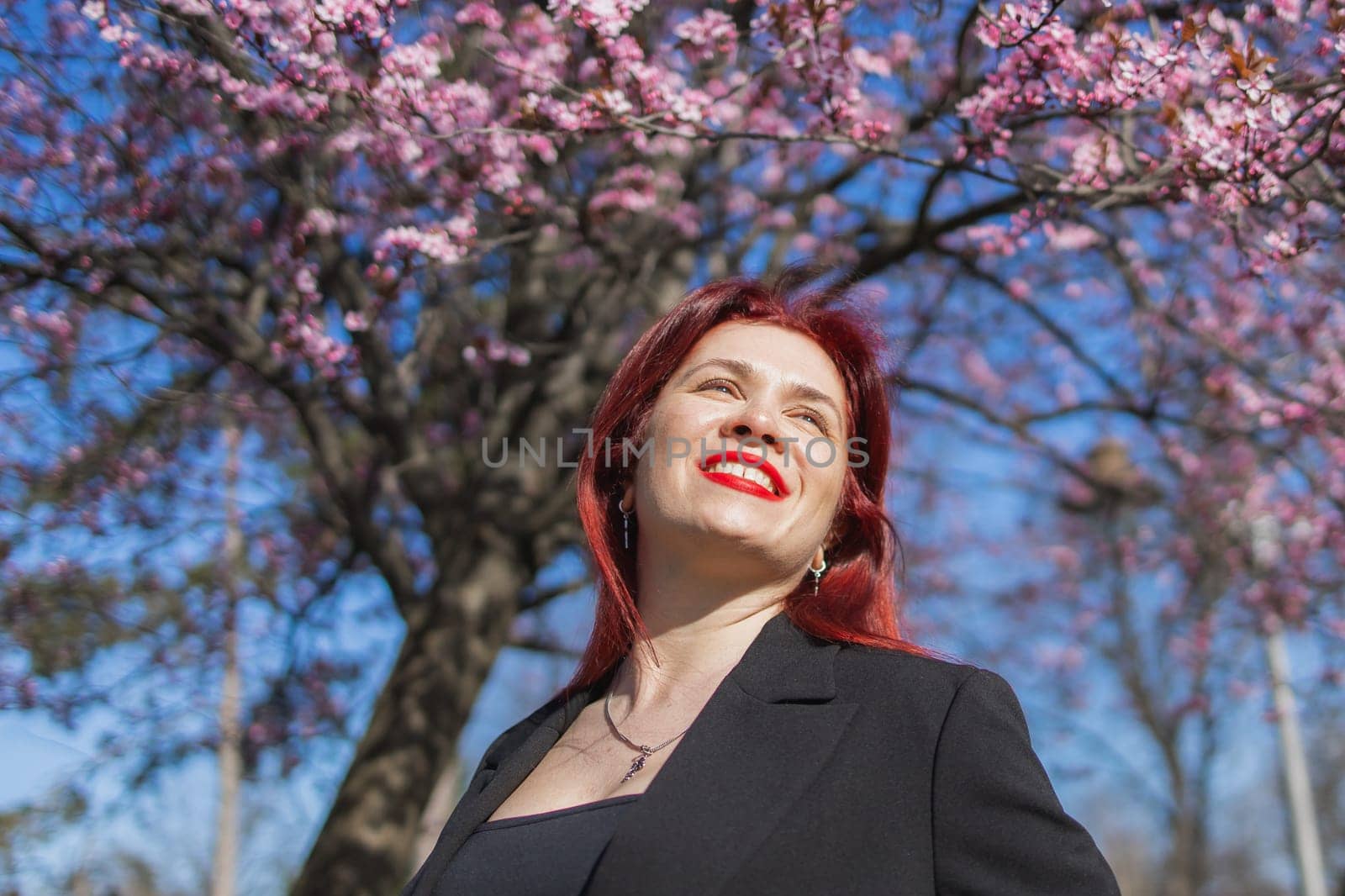 Fashion outdoor photo of beautiful woman with red curly hair in elegant suit posing in spring flowering park with blooming cherry tree. Copy space and empty place for advertising text.