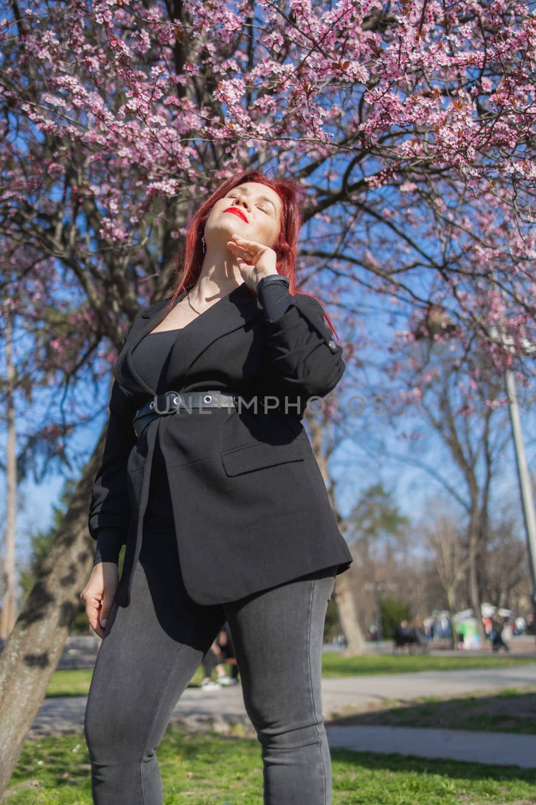 Fashion outdoor photo of beautiful woman with red curly hair in elegant suit posing in spring flowering park with blooming cherry tree. Copy space and empty place for advertising text by Satura86