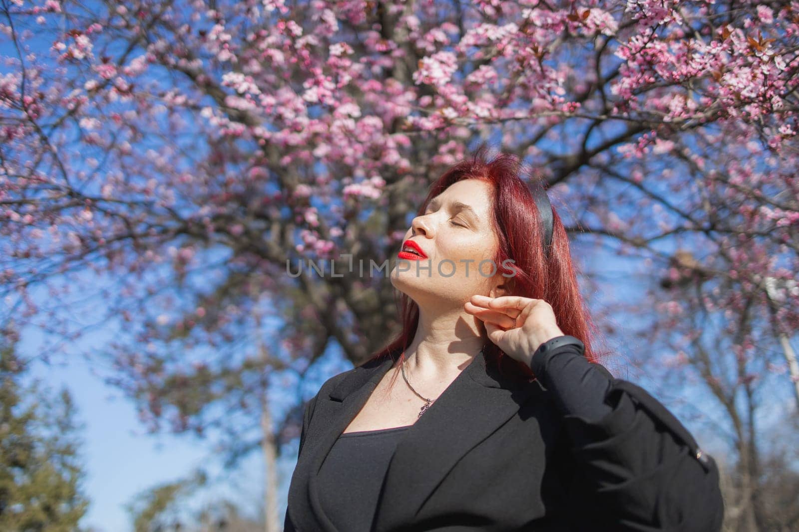 Fashion outdoor photo of beautiful woman with red curly hair in elegant suit posing in spring flowering park with blooming cherry tree. Copy space and empty place for advertising text by Satura86