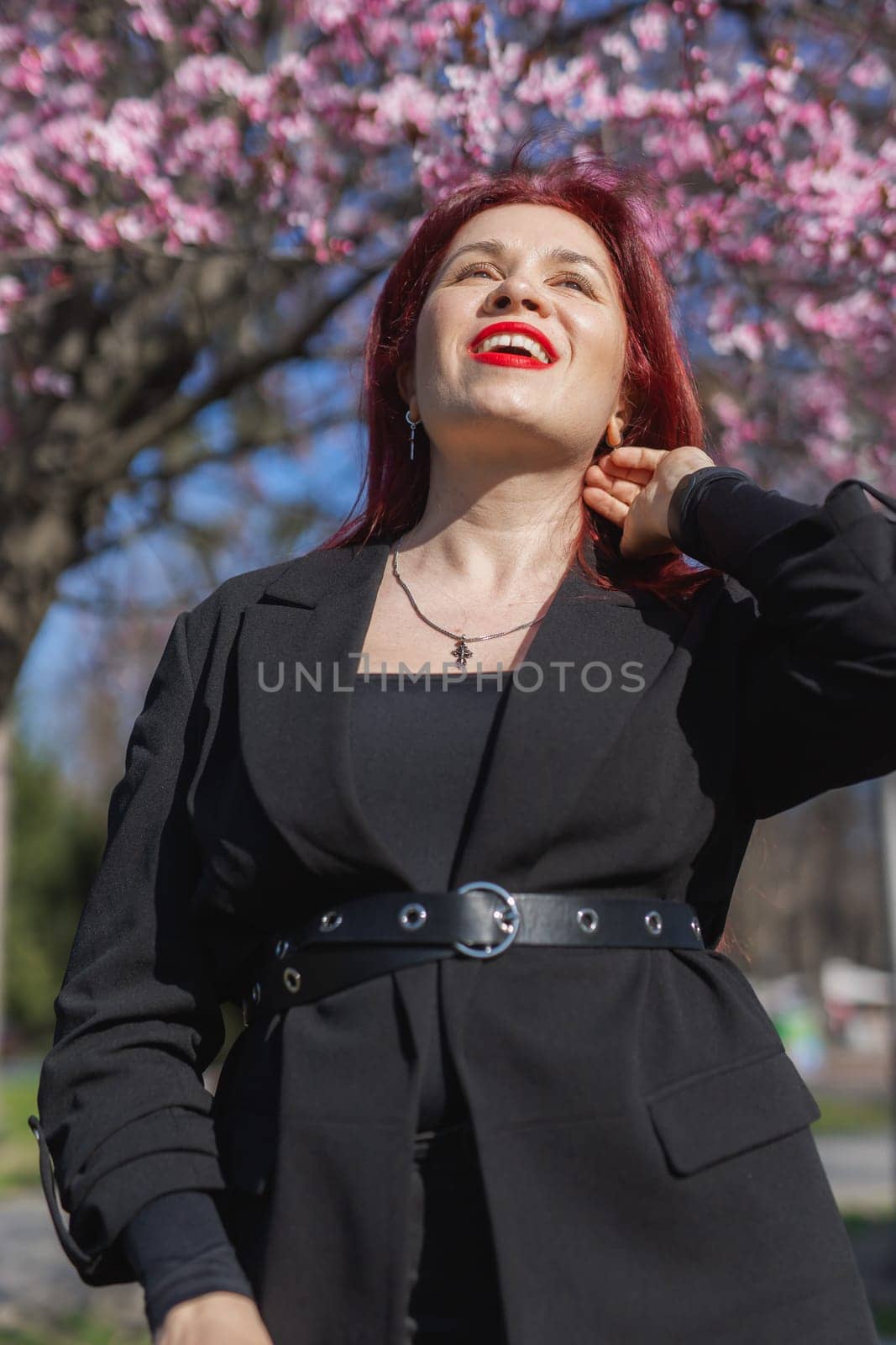 Fashion outdoor photo of beautiful woman with red curly hair in elegant suit posing in spring flowering park with blooming cherry tree. Copy space and empty place for advertising text by Satura86