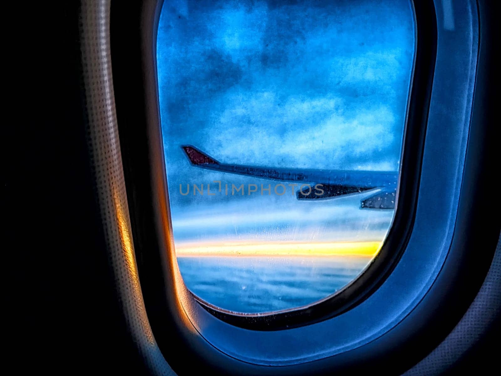 Airplane wing, clouds and sunrise through plane window. Wing silhouetted against vibrant sky and clouds. Sunset Horizon View From Airplane. Blurred