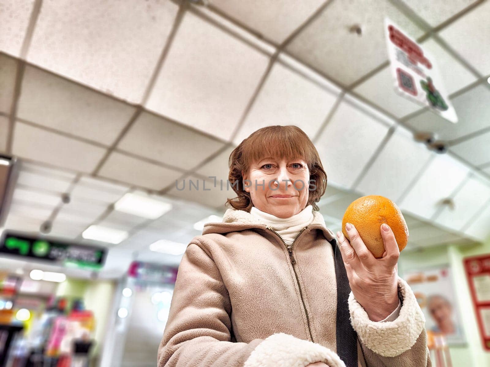 Middle-Aged Woman Holding an Orange in Grocery Store. Woman examines an orange, considering nutrition in a store aisle. Concept of proper nutrition