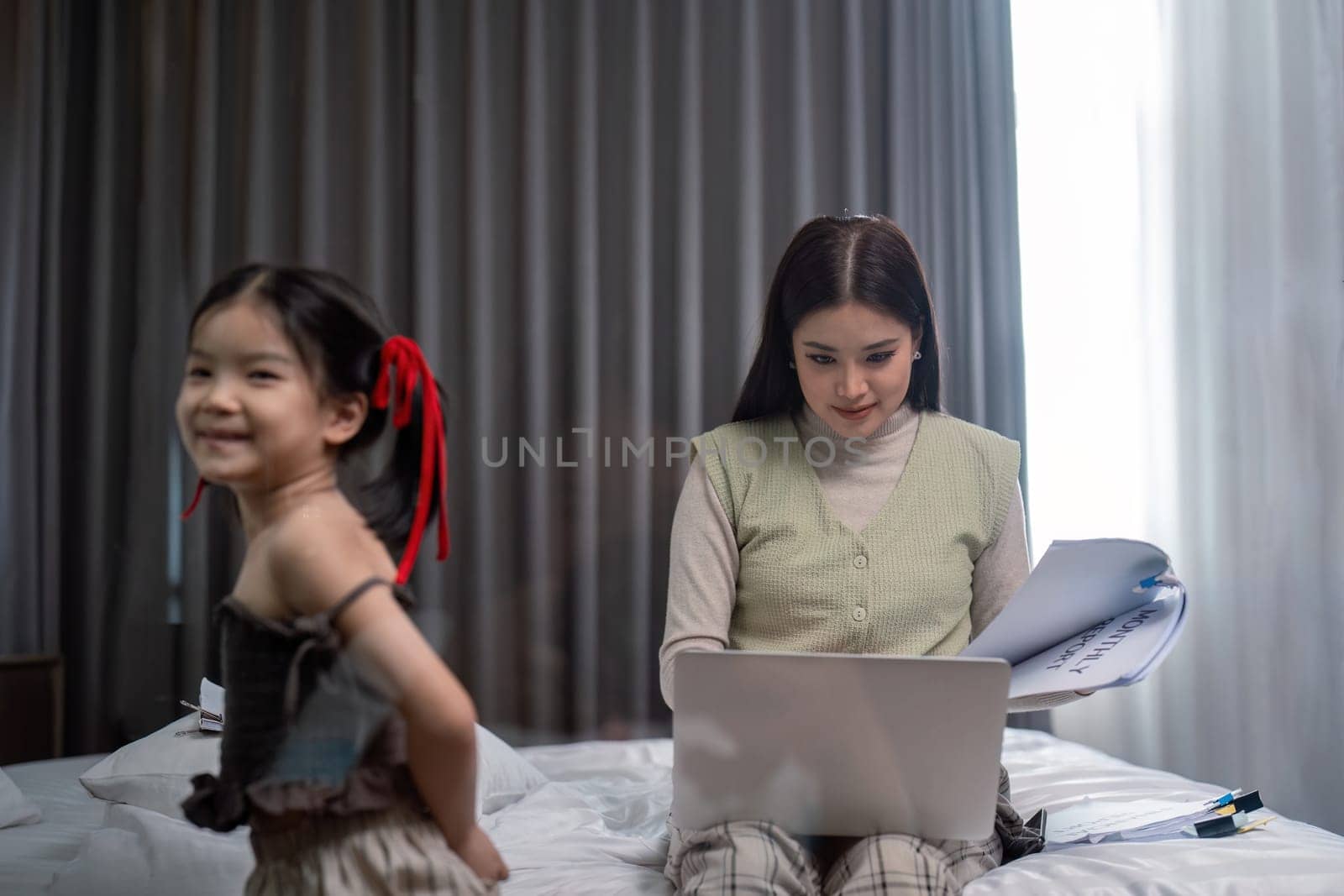 Mother and daughter lying in bed with on and smiling. Young mom working from home in bedroom with laptop.