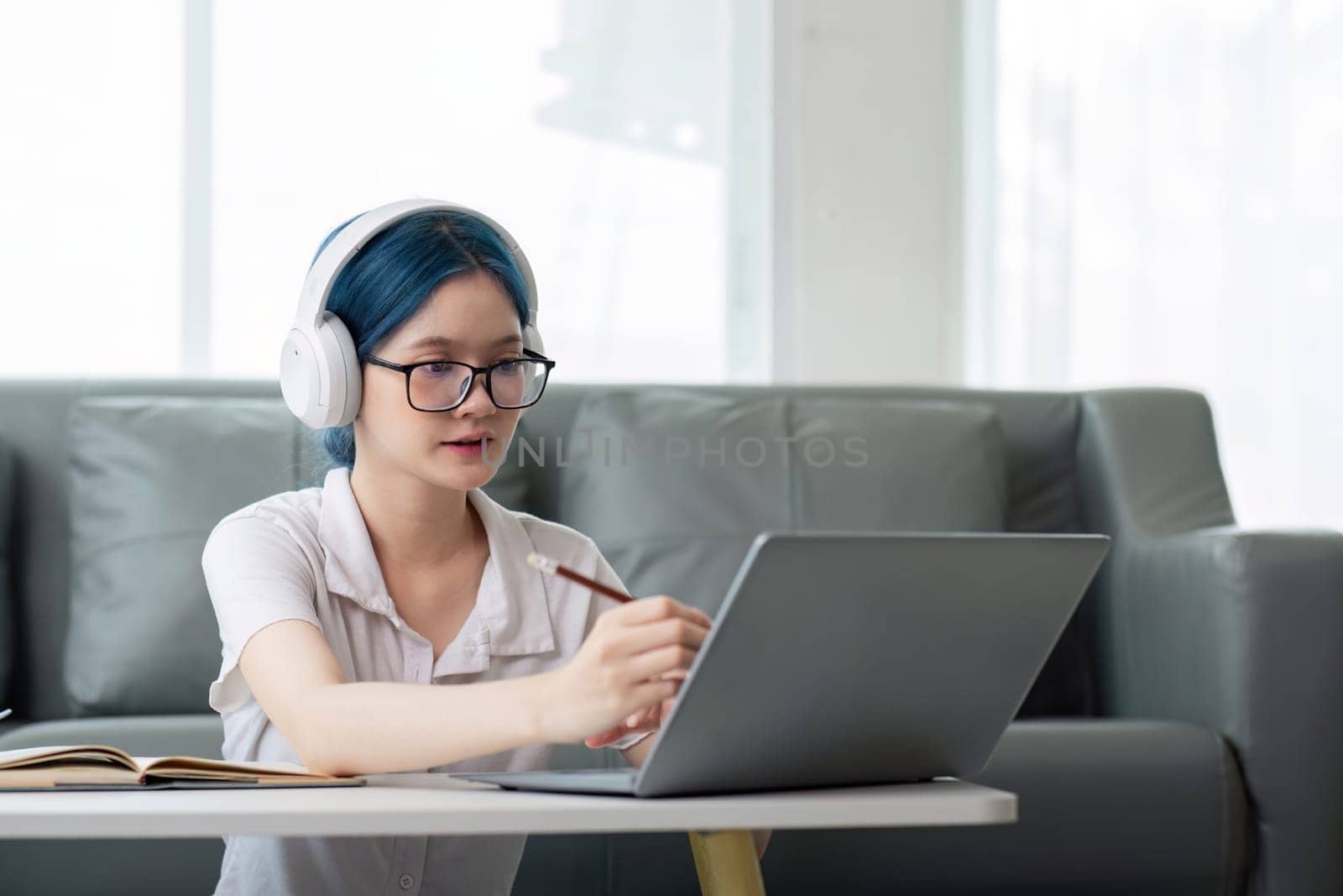 Smiling girl student wear headphone study online with teacher, happy young asian woman learn lecture watch webinar write notes look at laptop sit in home, distant education.