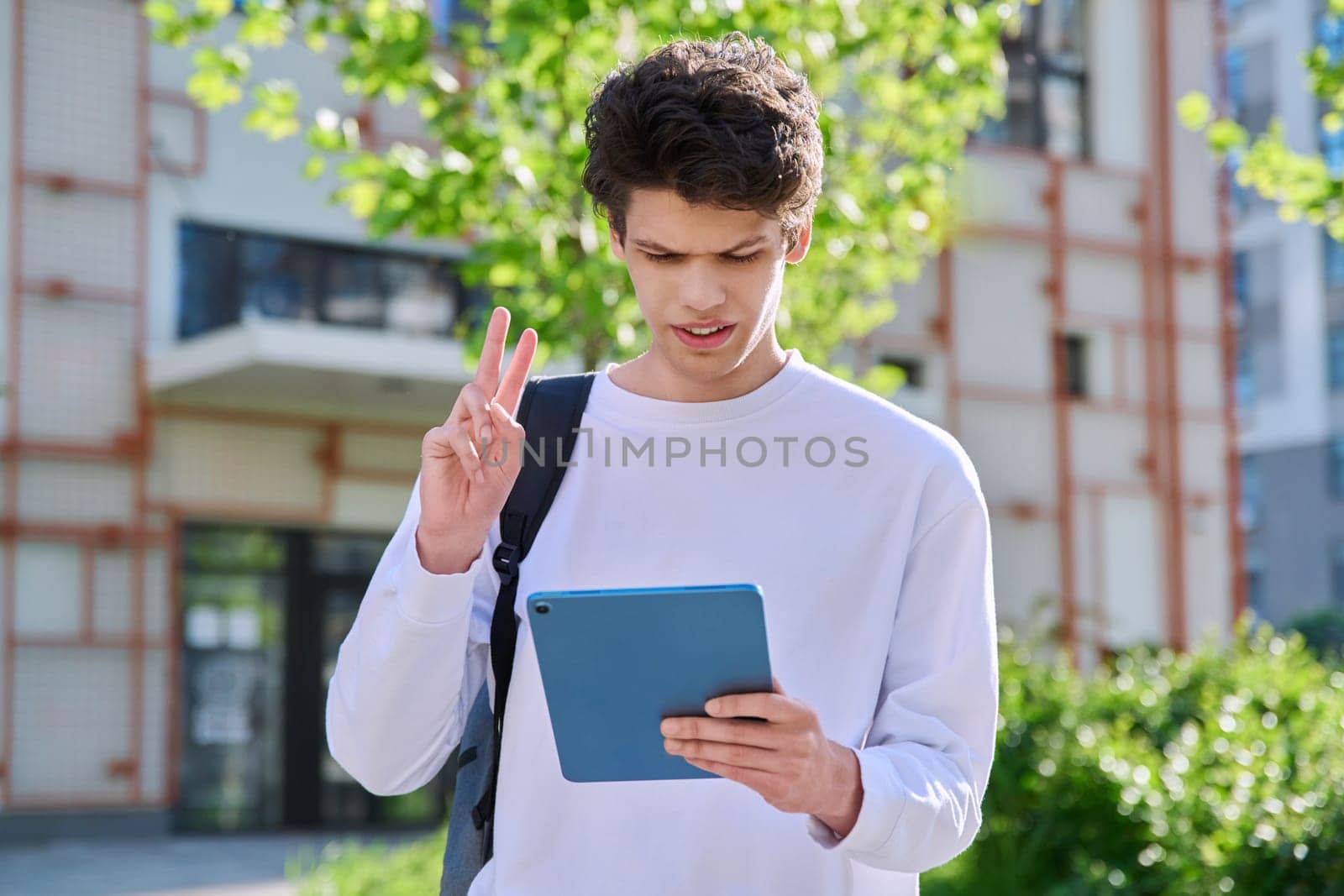 Young handsome guy college student using digital tablet outdoor, talking on video call chat conference, educational building background. Education, technology, training, 19,20 years age youth concept